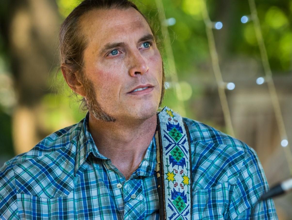 Man in blue plaid shirt outdoors, gazing thoughtfully.
