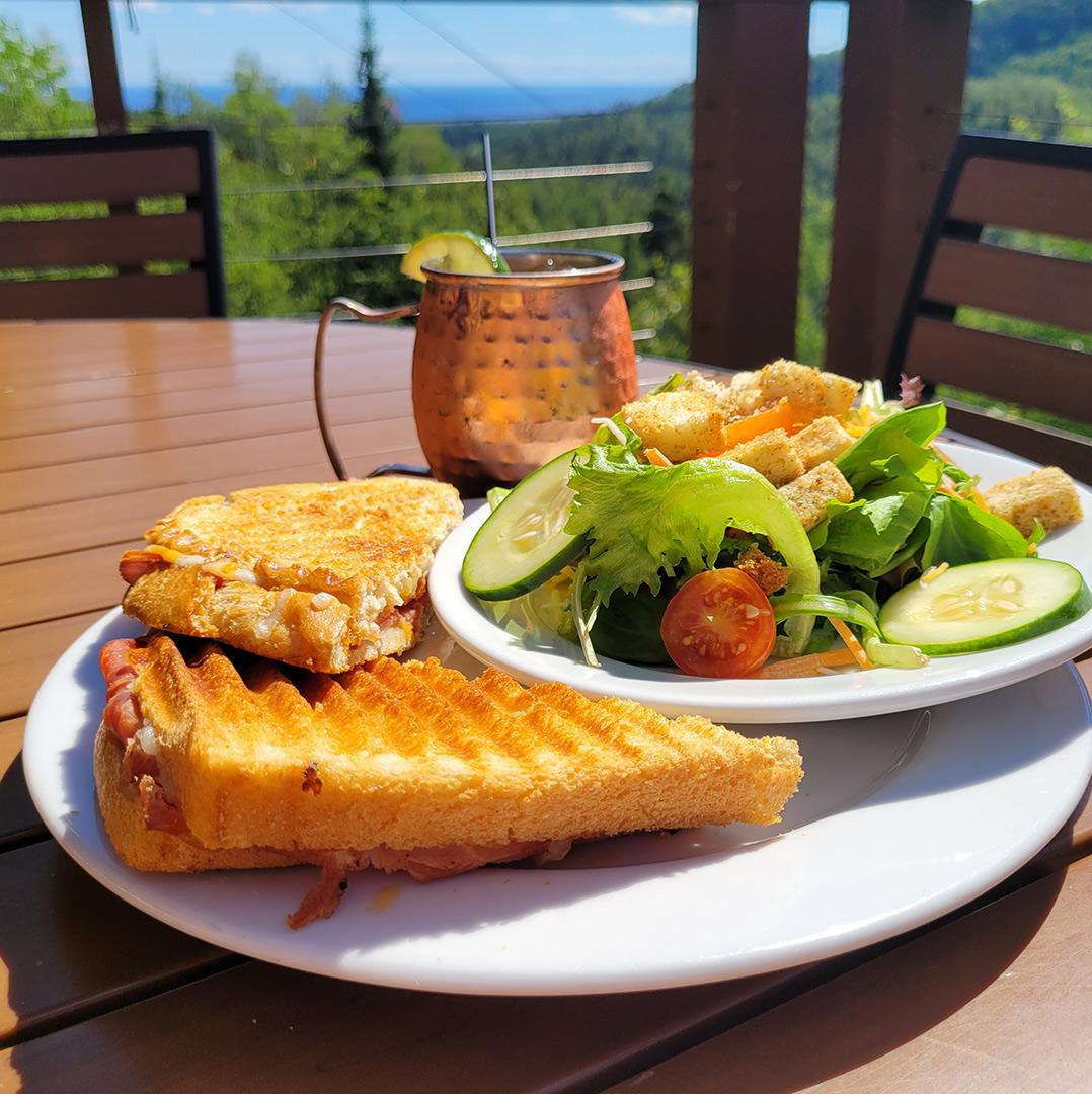 Grilled sandwich, salad, and drink on table with mountain view.