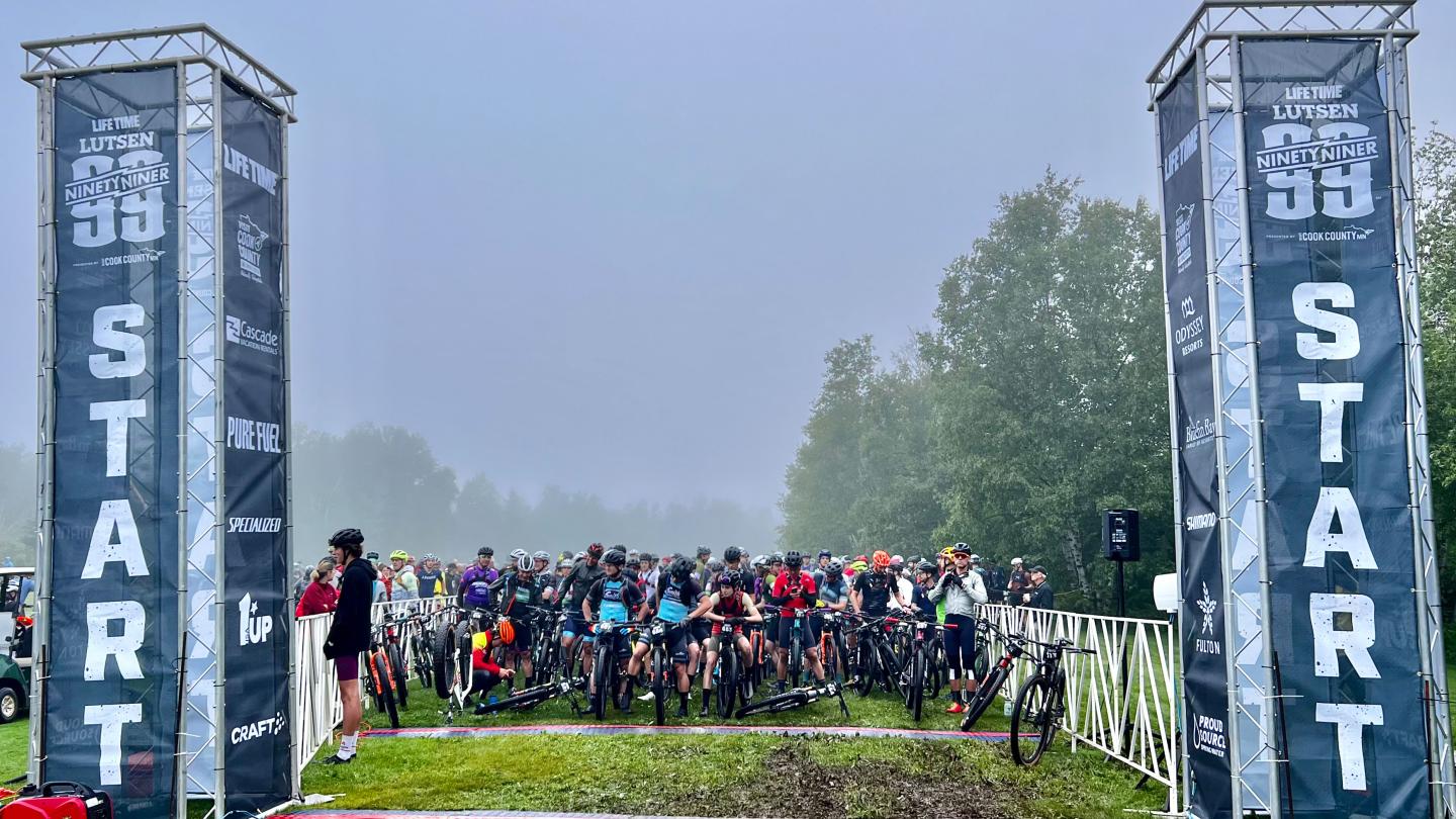 Cyclists gathered at a foggy race start line between two tall banners.
