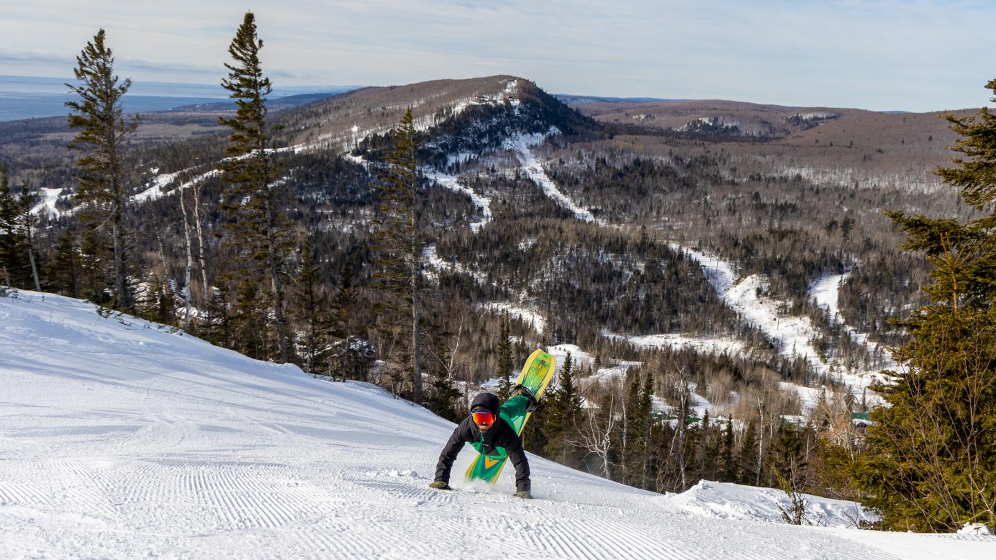Snowboarder descending a snowy mountain with forested hills in the background.