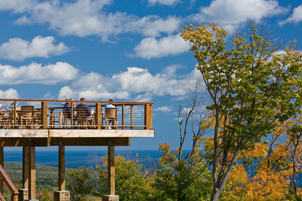 Wooden deck with people overlooking a scenic view, trees, and a bright blue sky.