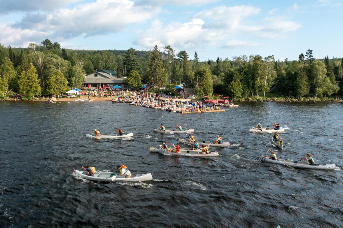 Canoes racing on a river, with a crowd onshore and trees in the background.