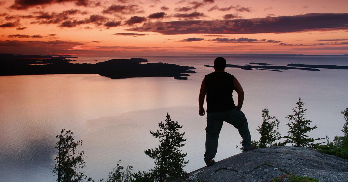 Silhouette of person on cliff overlooking lake at sunset.