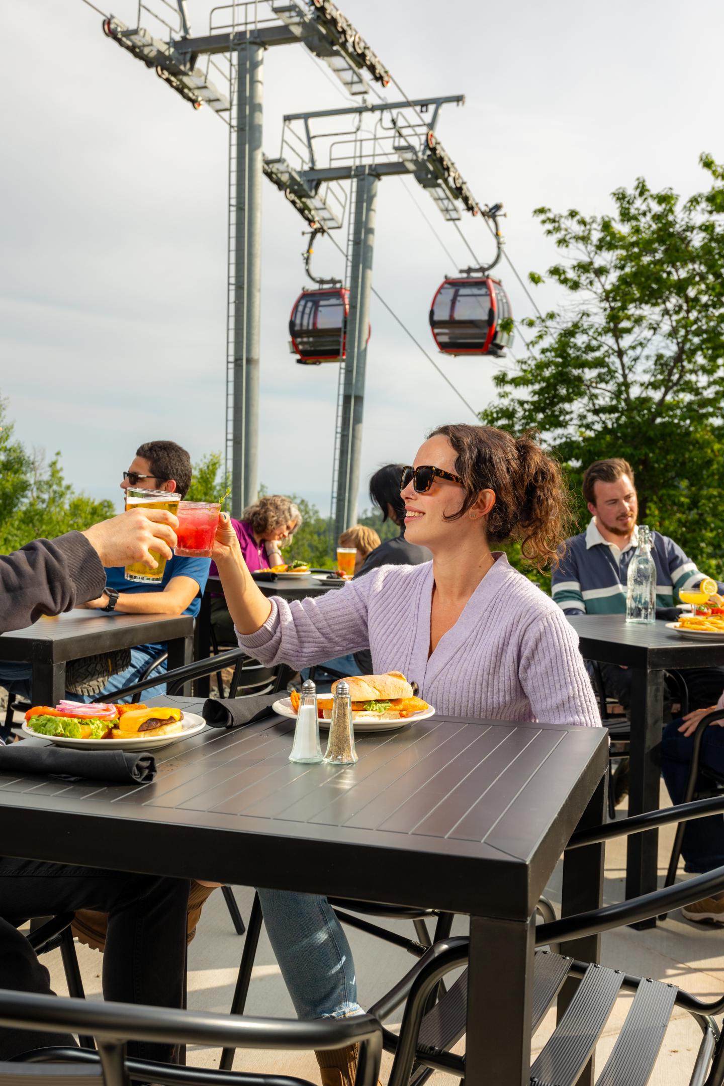 Outdoor dining with people toasting drinks, aerial cable cars in background.