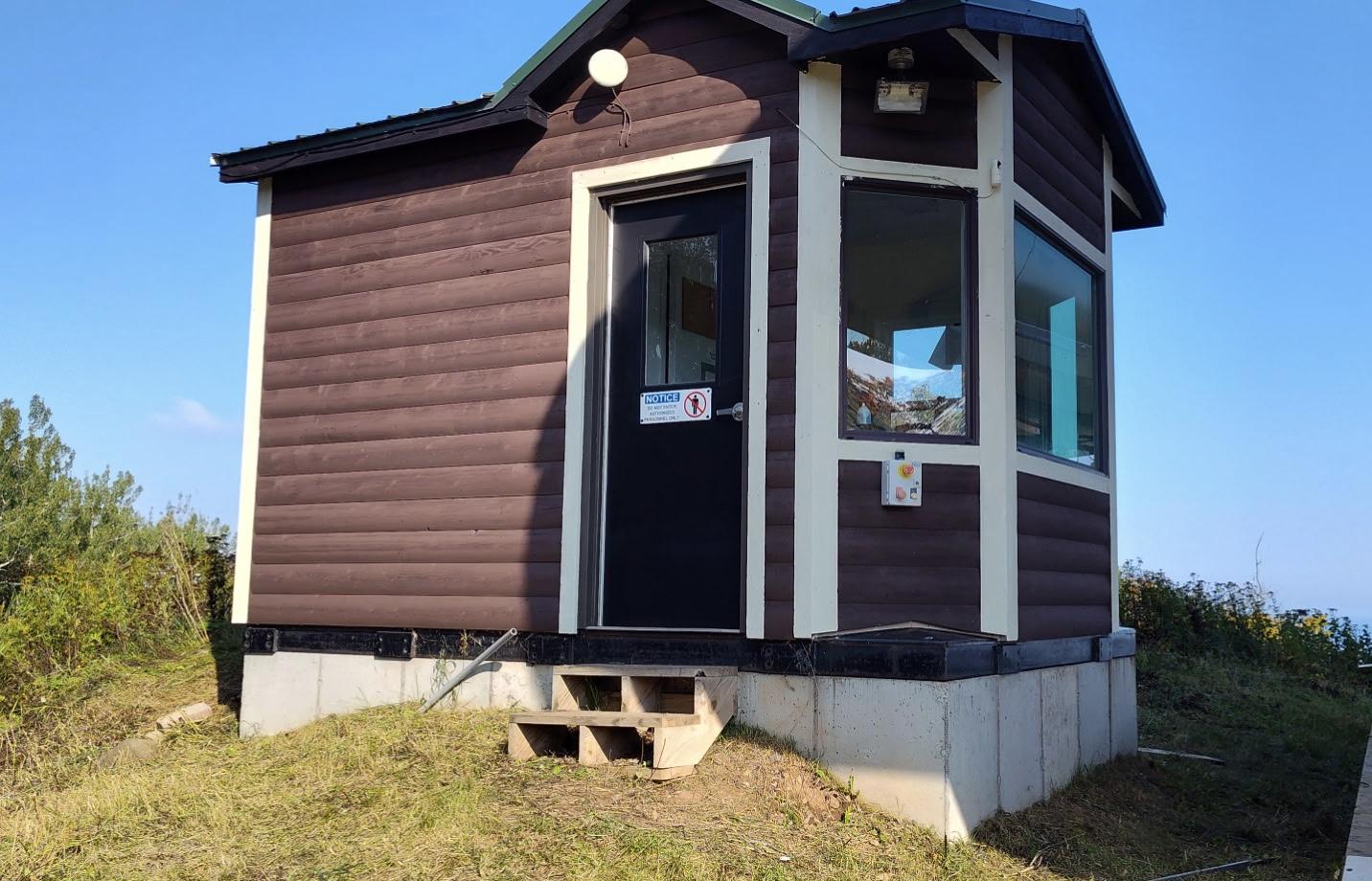 Small brown cabin with a black door on a grassy hill, under blue sky.