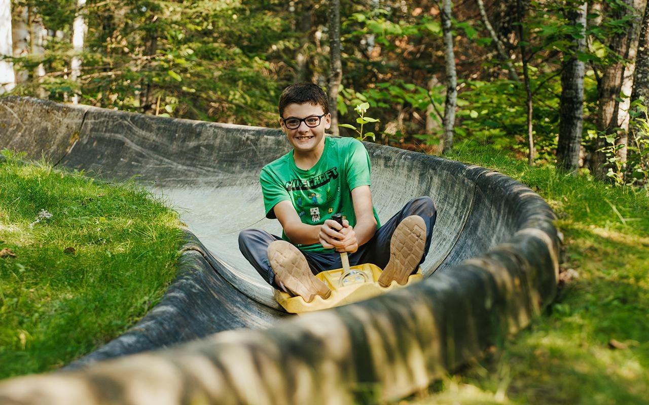 Child smiling while riding a slide in a forest setting.