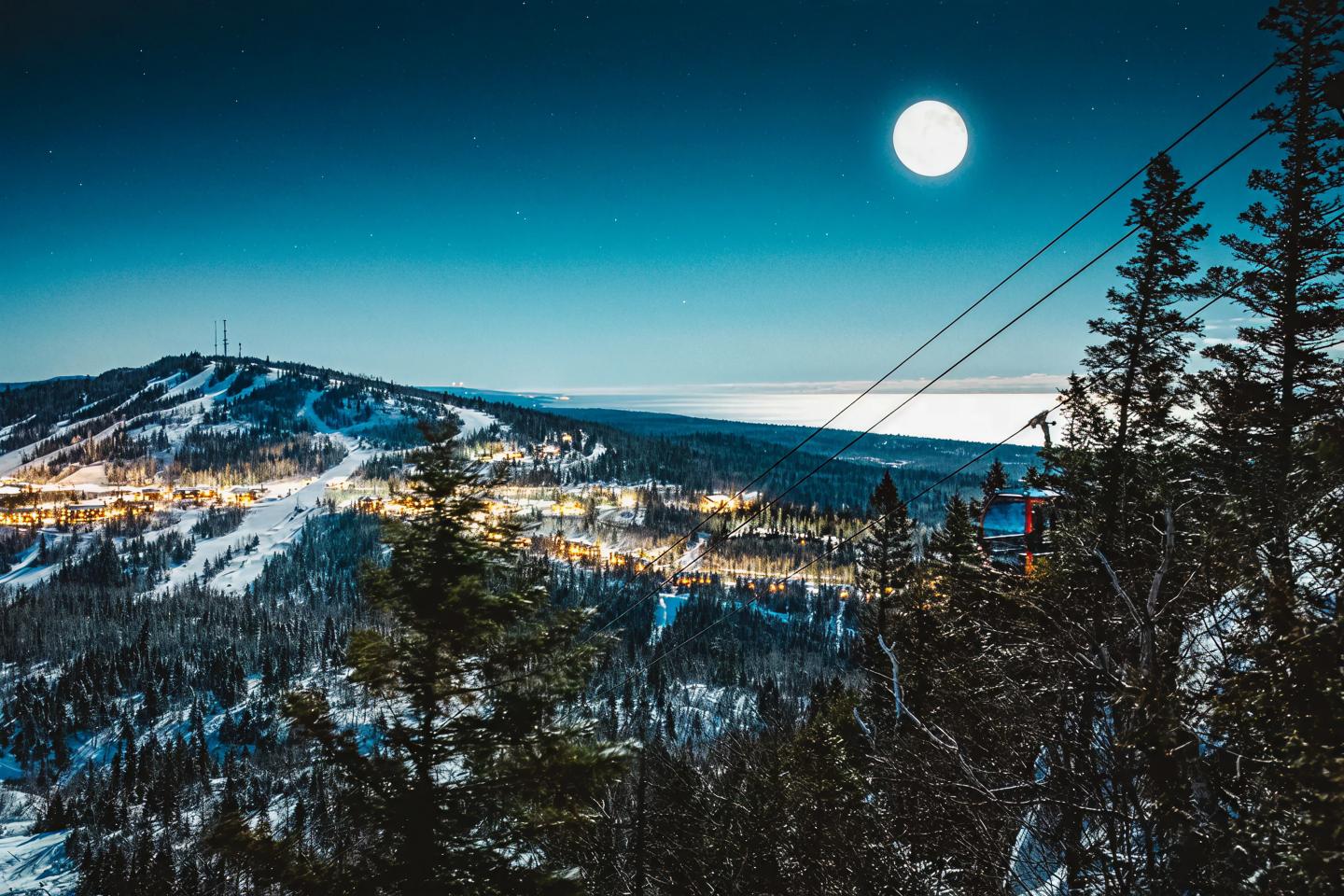 Snowy mountain landscape under a full moon, with city lights in the distance.