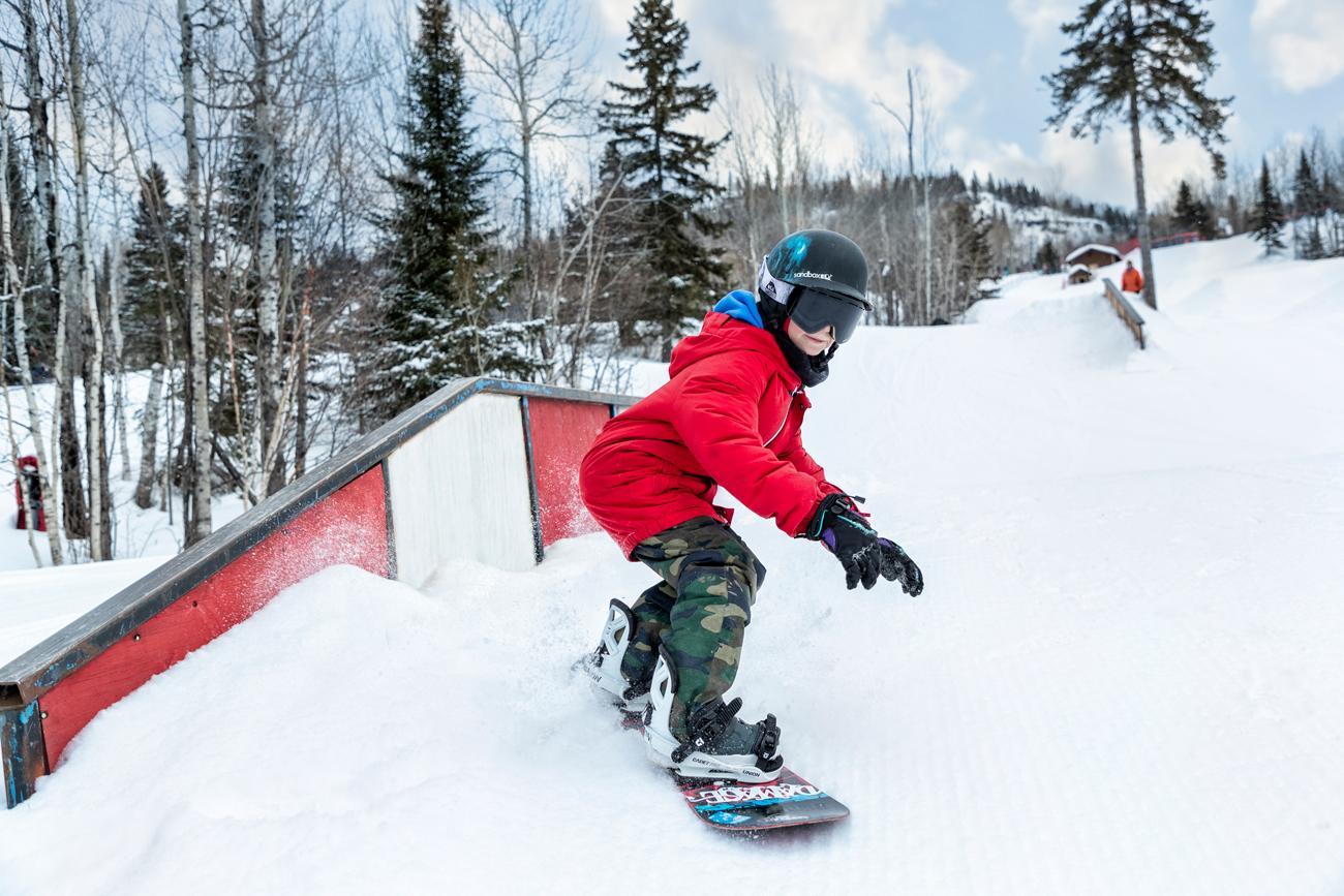 Snowboarder in red jacket riding down a snowy slope, surrounded by trees.