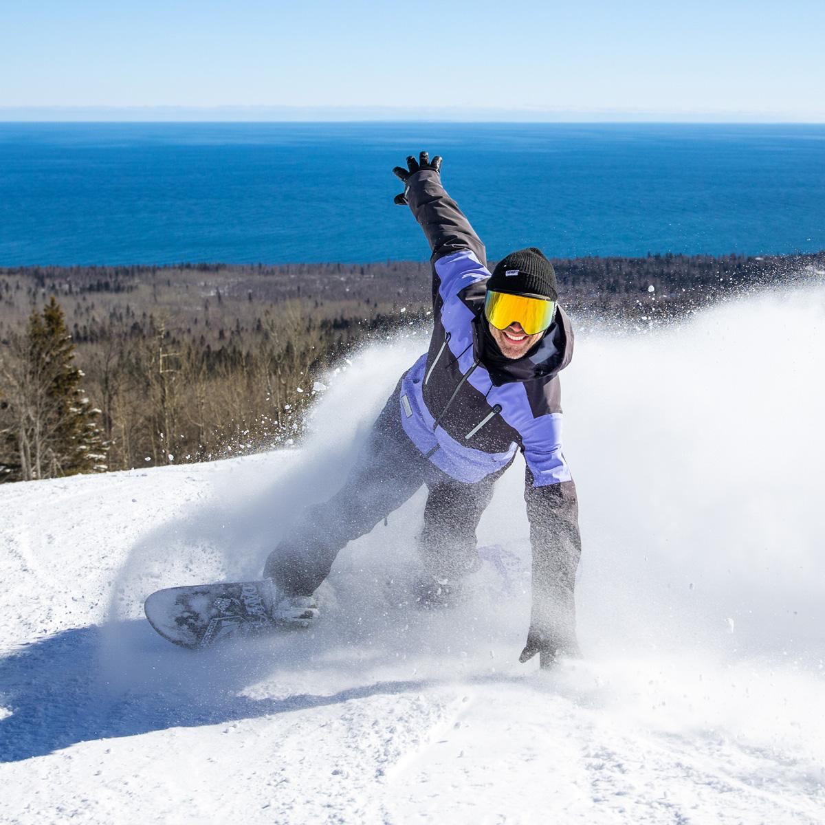 Snowboarder carving down a snowy slope with ocean view.