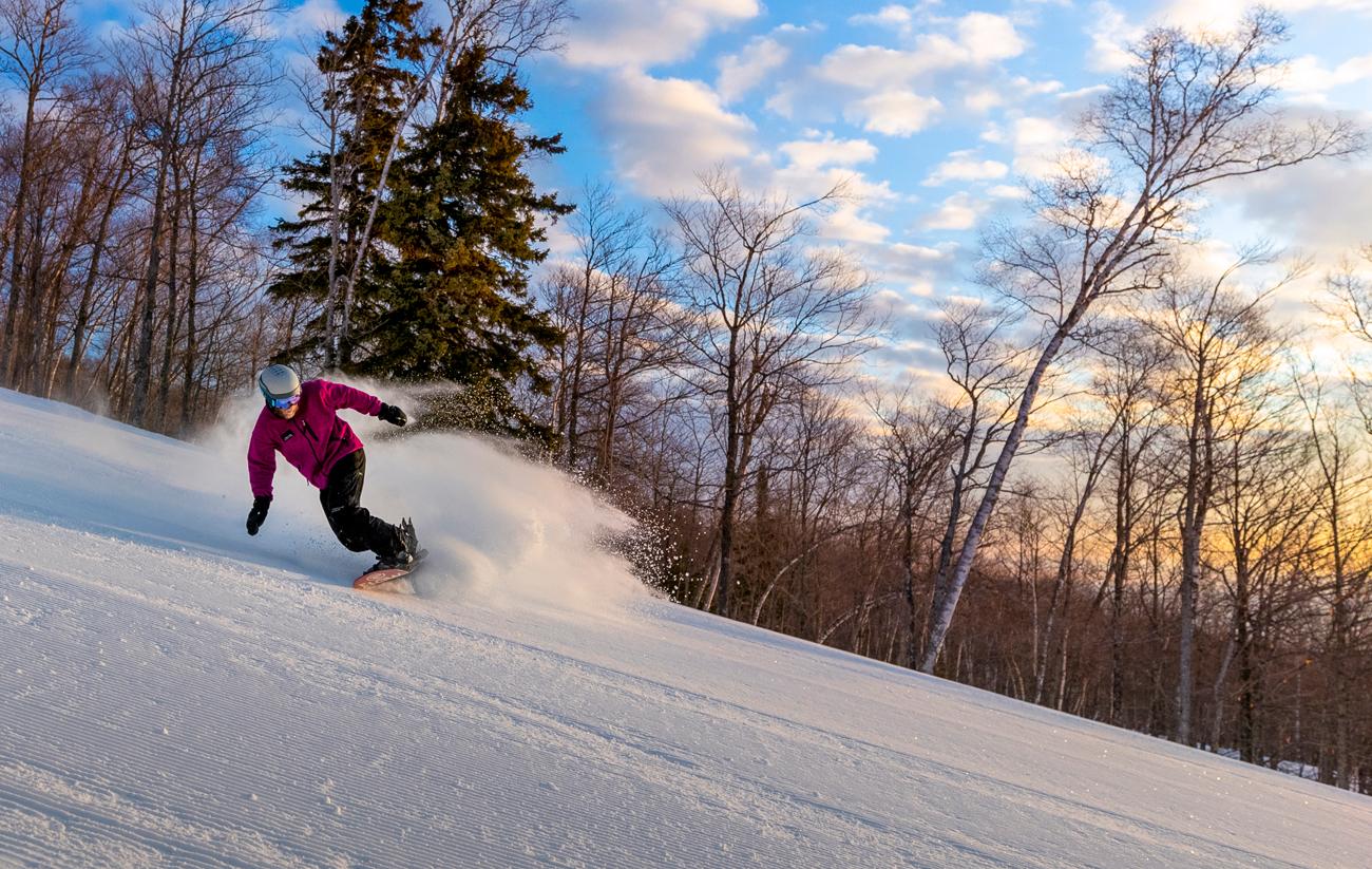 Snowboarder gliding downhill at sunset, surrounded by trees.
