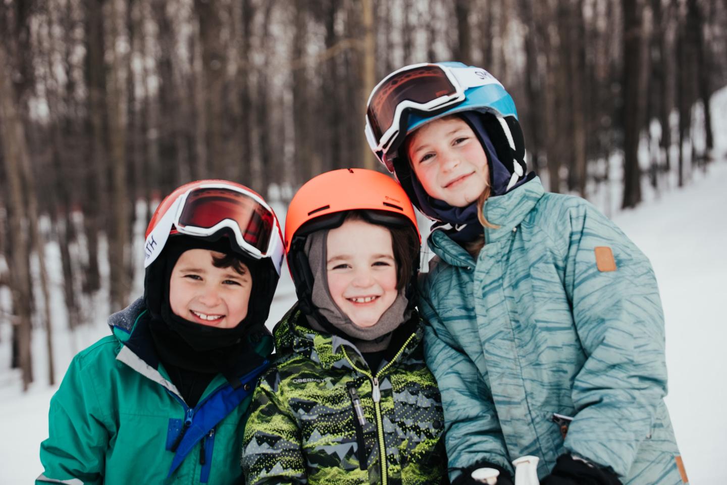 Three smiling kids in winter gear and helmets, standing in a snowy forest.
