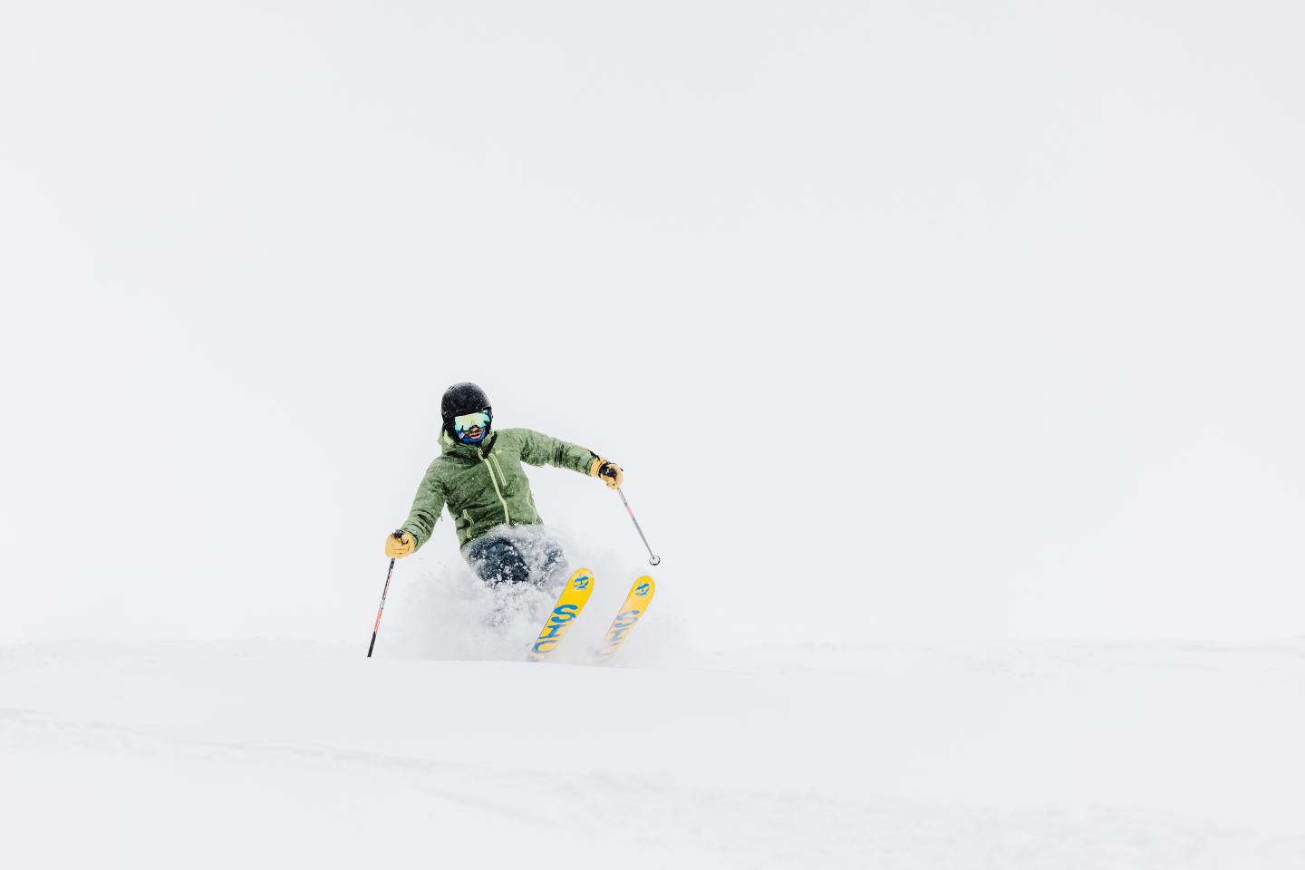 Skier in green jacket descending a snowy slope against a white backdrop.