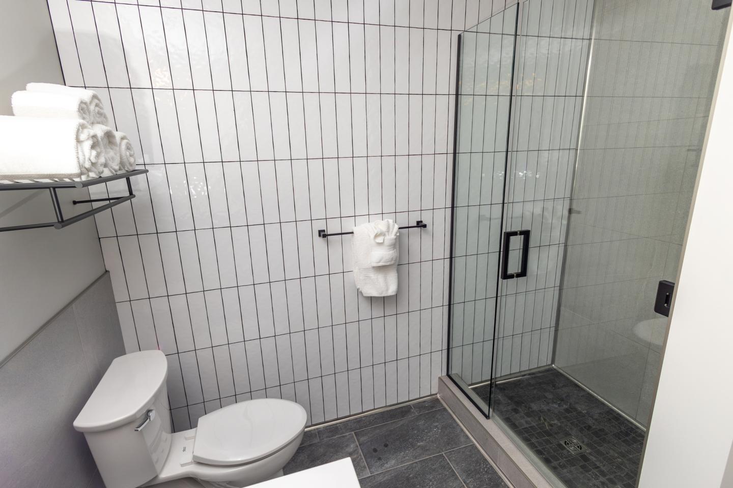 Bathroom with a glass shower, toilet, and shelves with rolled towels.