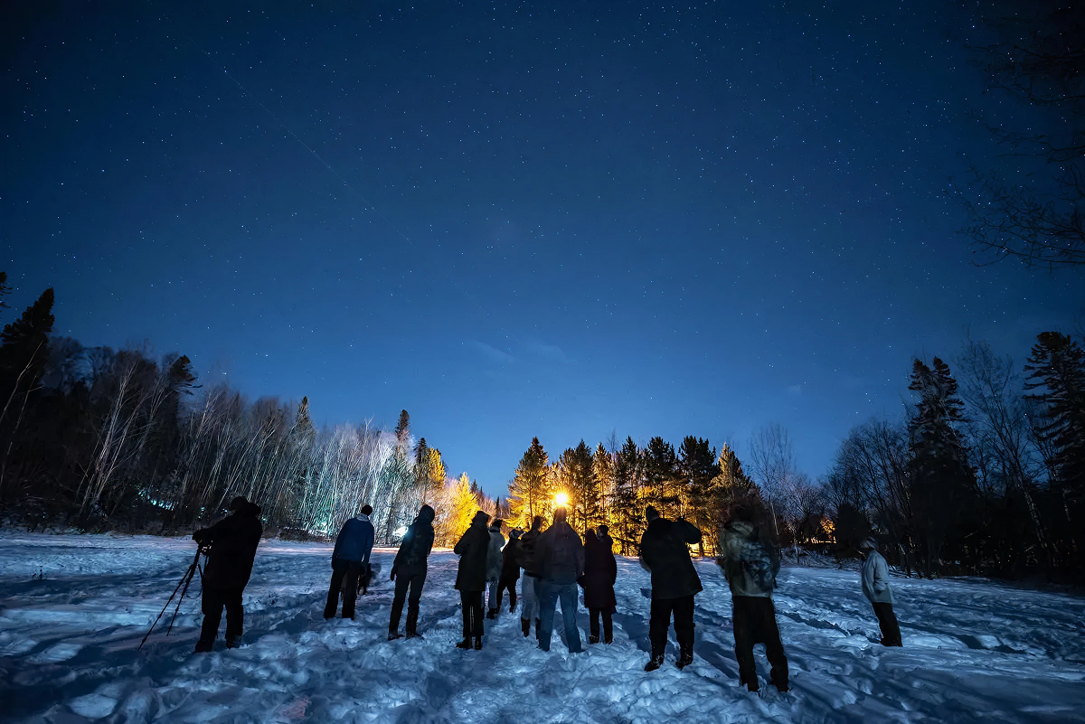 Night sky with stars, people stargazing in snowy forest clearing.