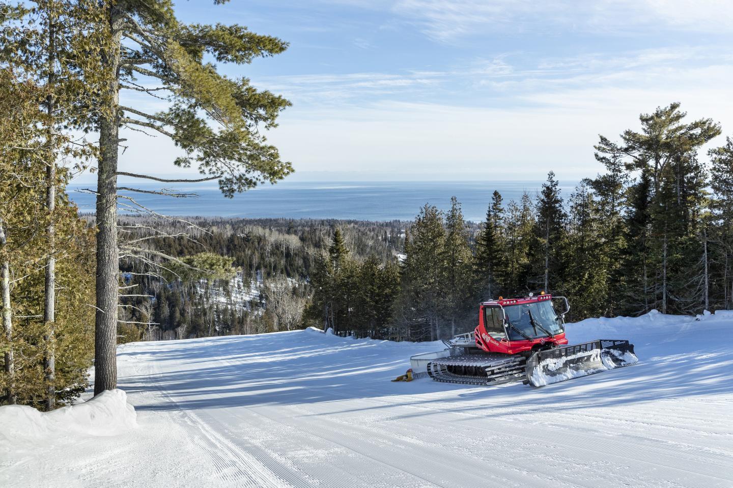 Red snow groomer on a snowy mountain slope, with trees and distant view of water.