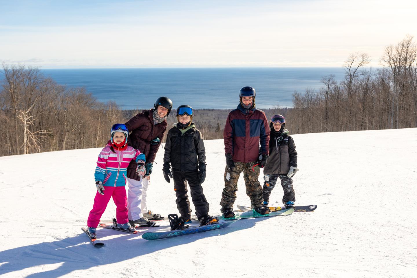 Group of five snowboarders on a snowy hill with ocean view.