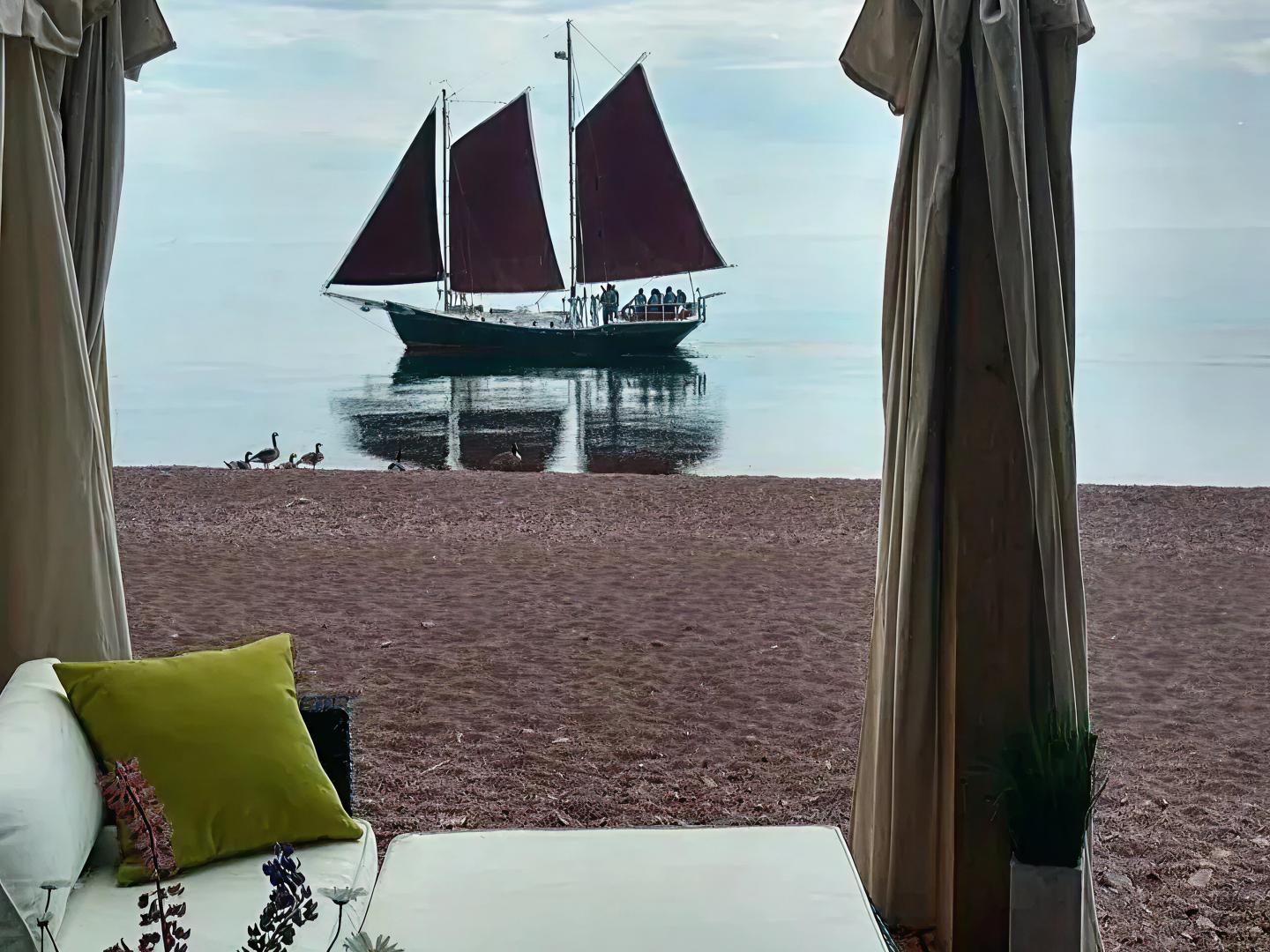 Sailboat with red sails on calm water, viewed from a cozy seaside cabana.