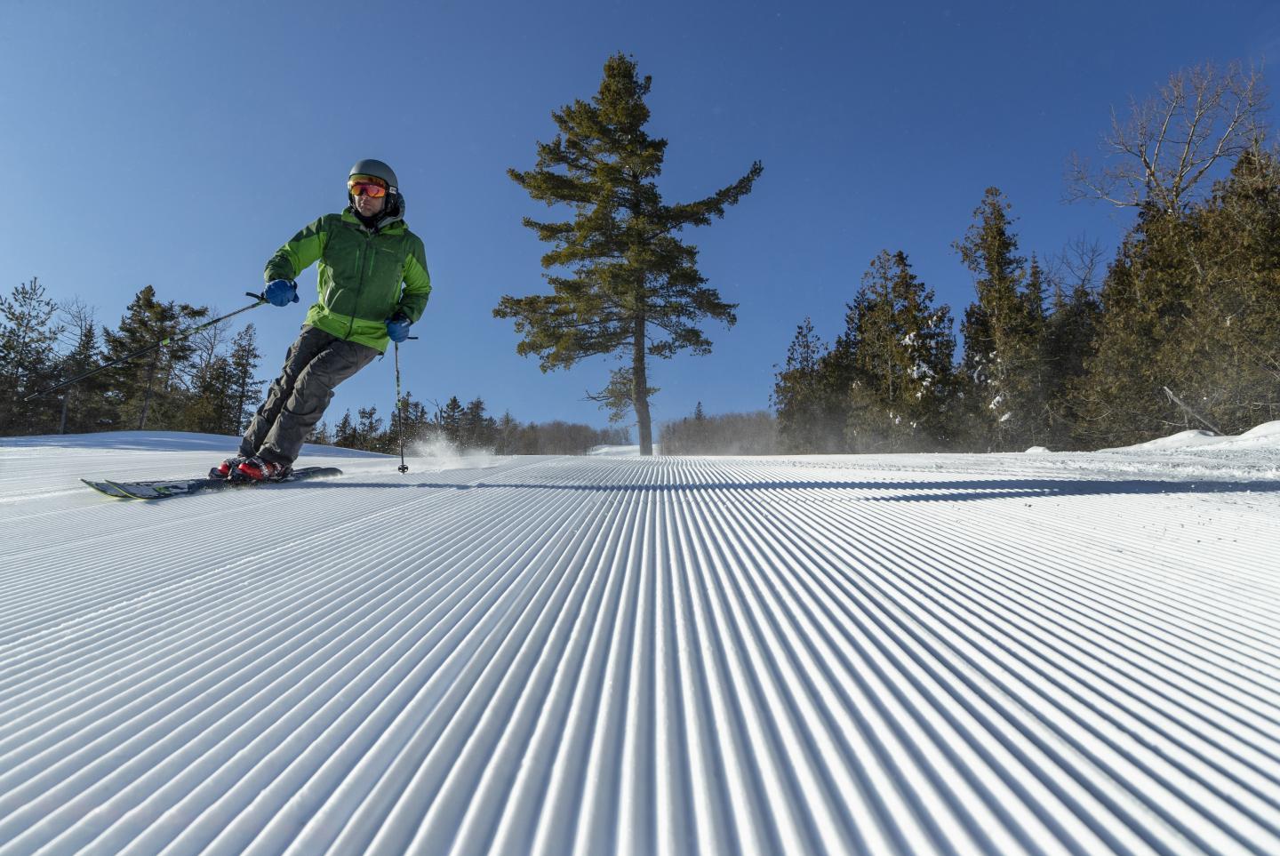 Skier in green jacket glides downhill on groomed snowy slope under a clear blue sky.