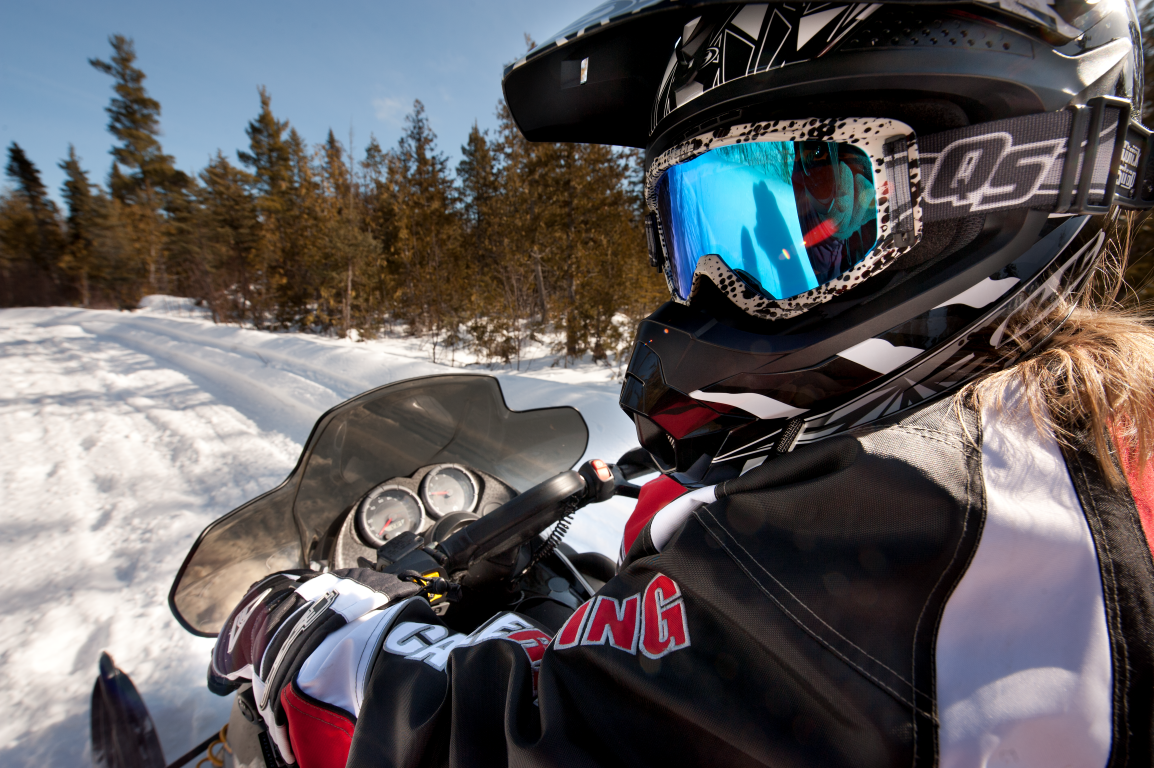 Snowmobiler wearing goggles and helmet on a snowy trail lined with trees.