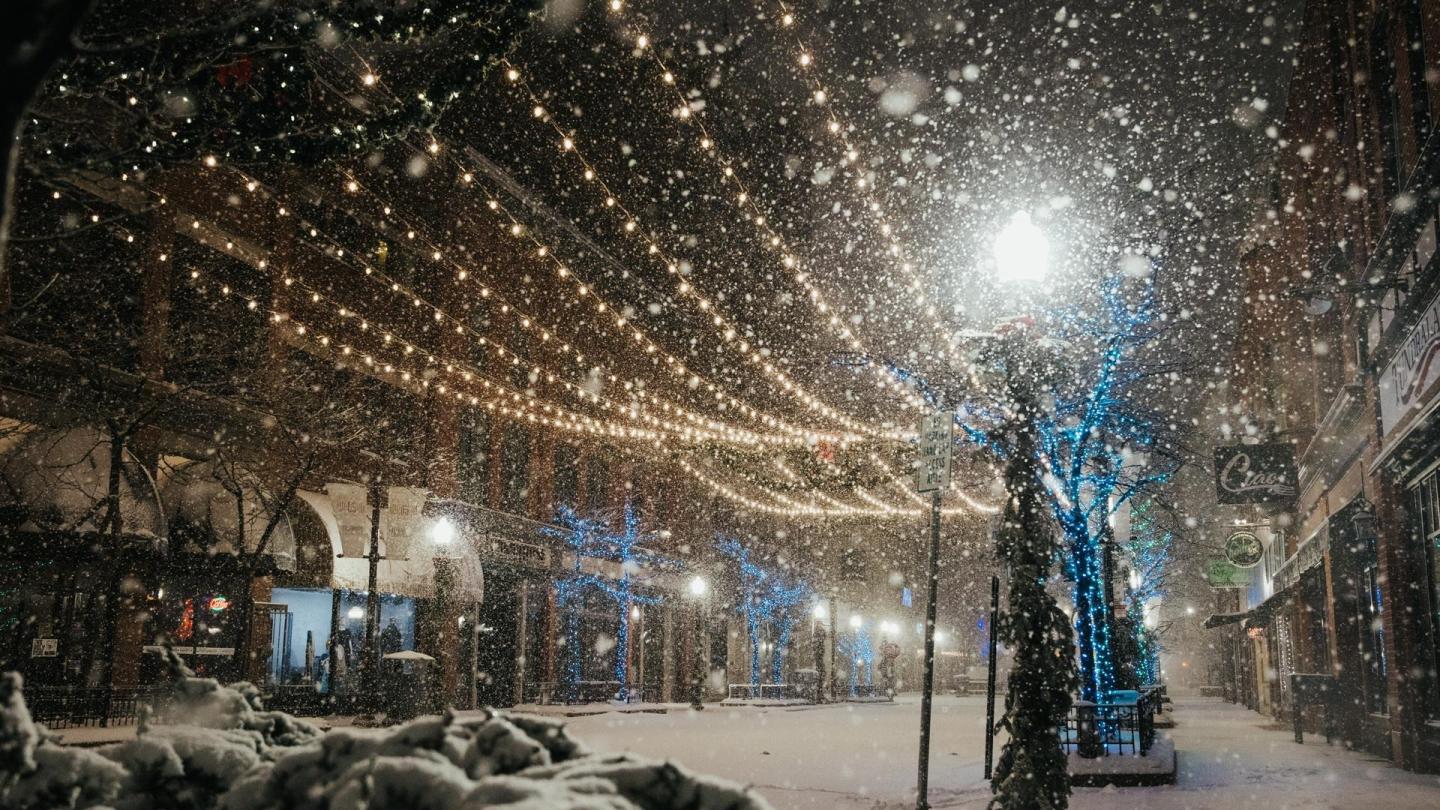 Snowy street with string lights and blue-lit trees.