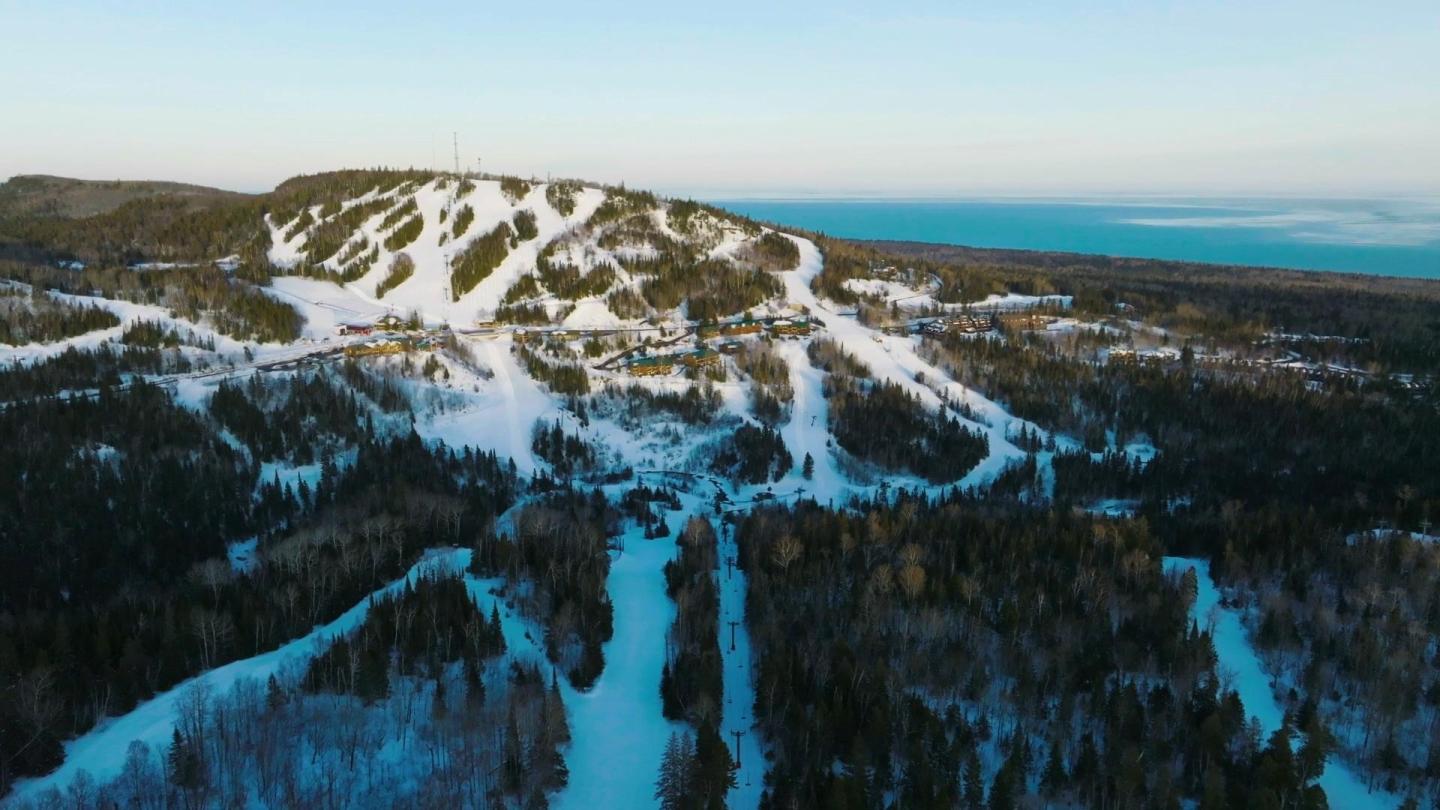 Snow-covered mountain with ski trails, surrounded by forest, under a clear blue sky.