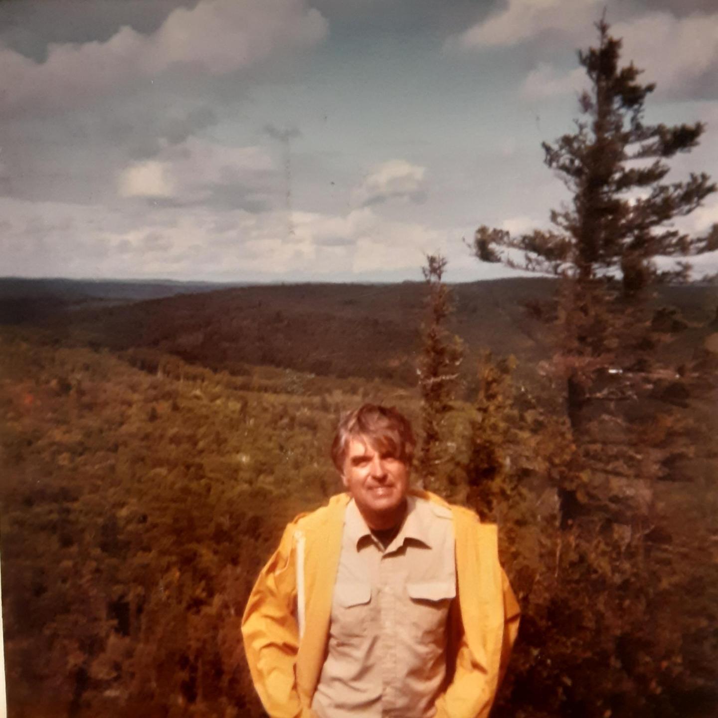 Man in a yellow jacket stands in front of a forested landscape.