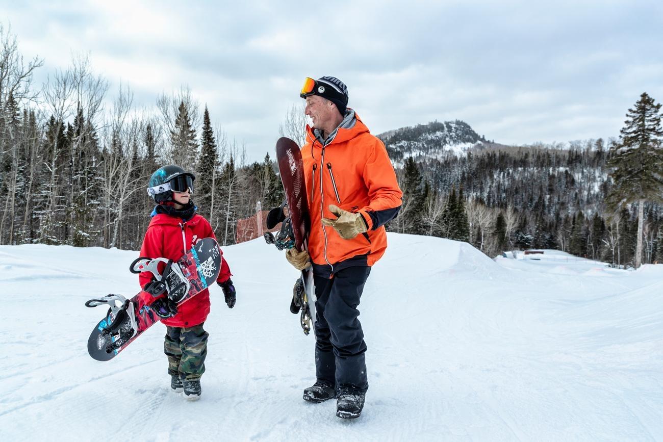 Child and adult with snowboards chatting on a snowy mountainside.