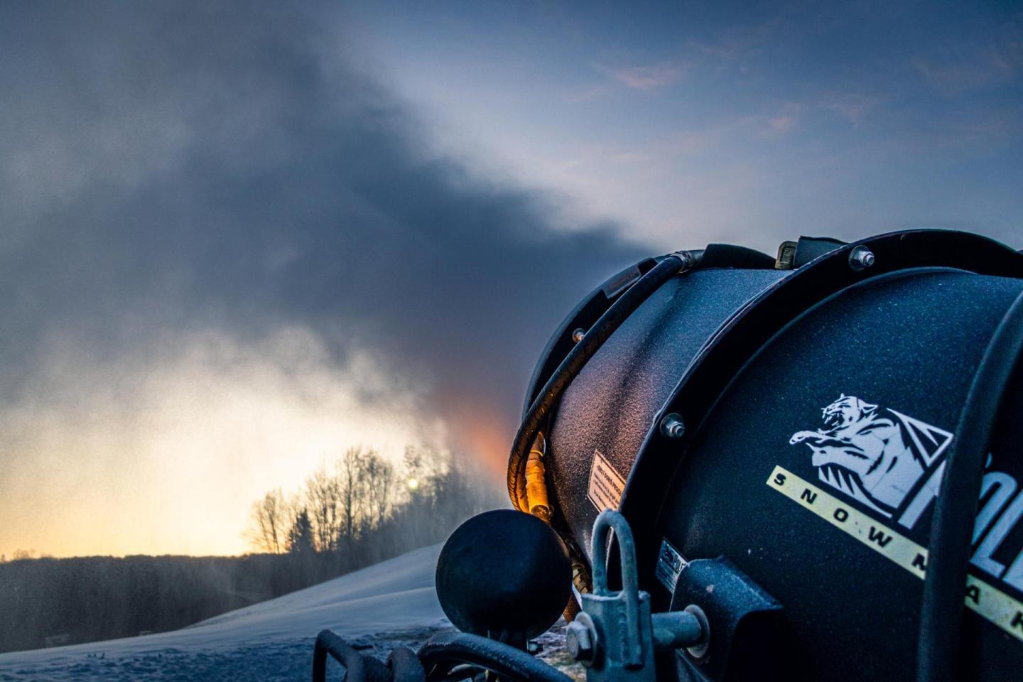 Snow cannon in sunset, mist rising, silhouetted trees in the background.