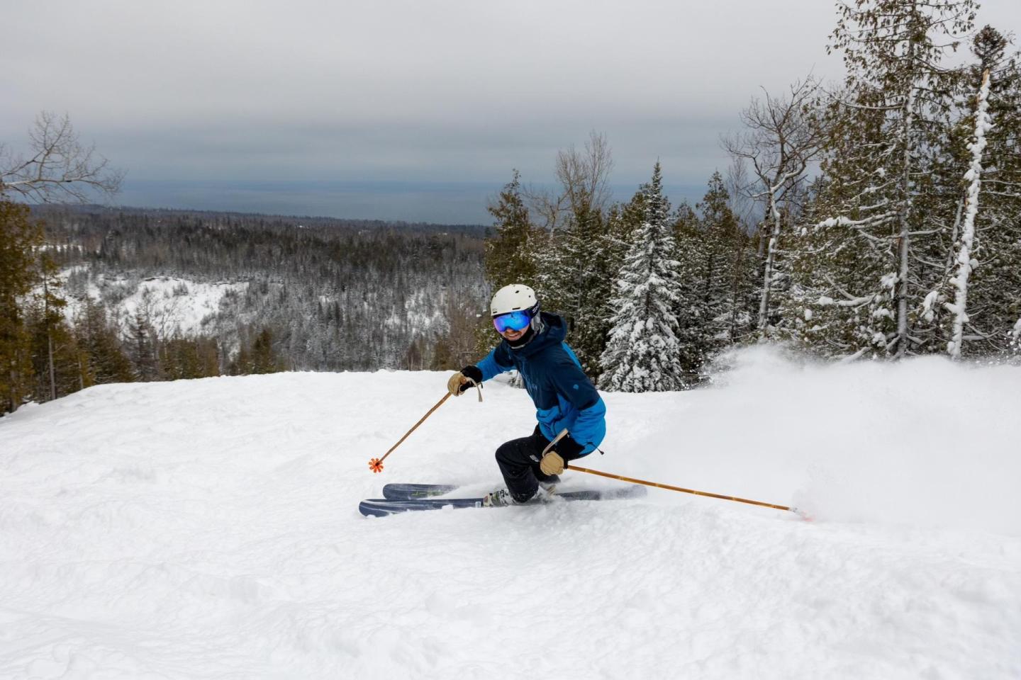 Skier in blue jacket descends snowy slope, surrounded by trees.
