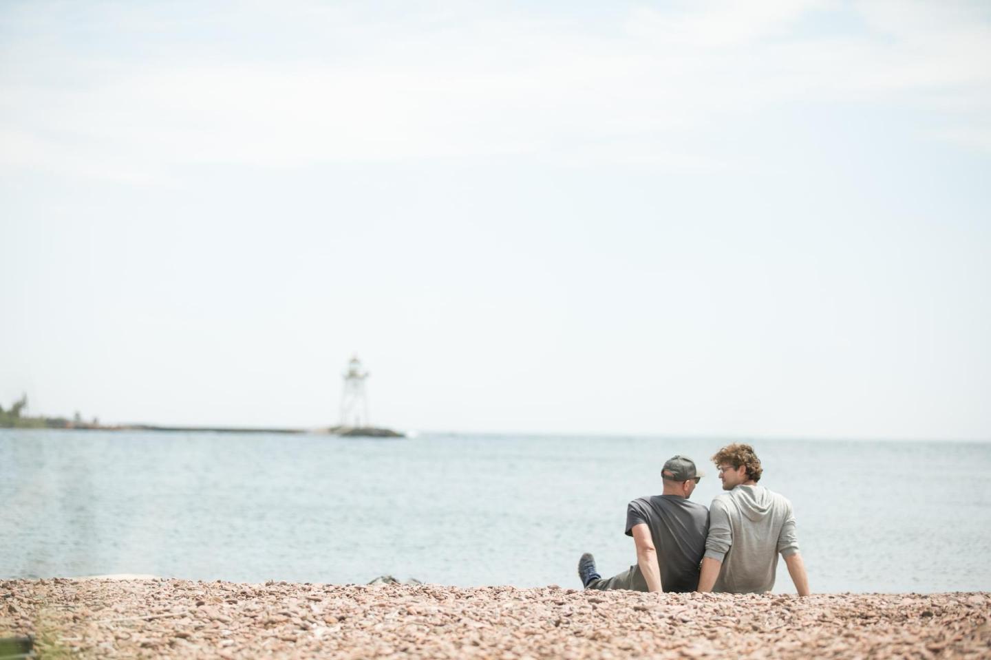 Two people sitting on a beach looking at the sea and distant lighthouse.