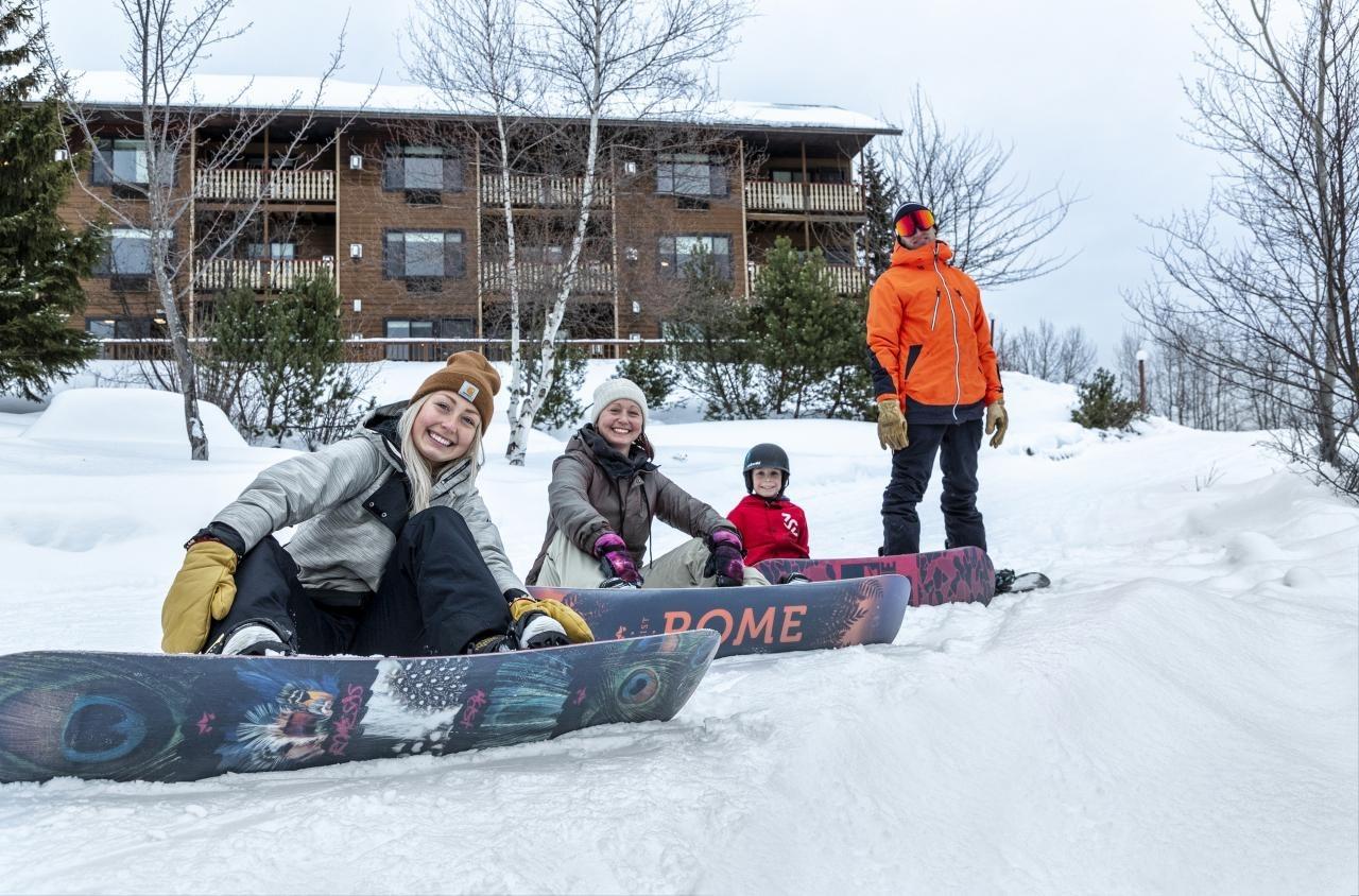 Snowboarders smiling on a snowy slope, with a wooden lodge in the background.
