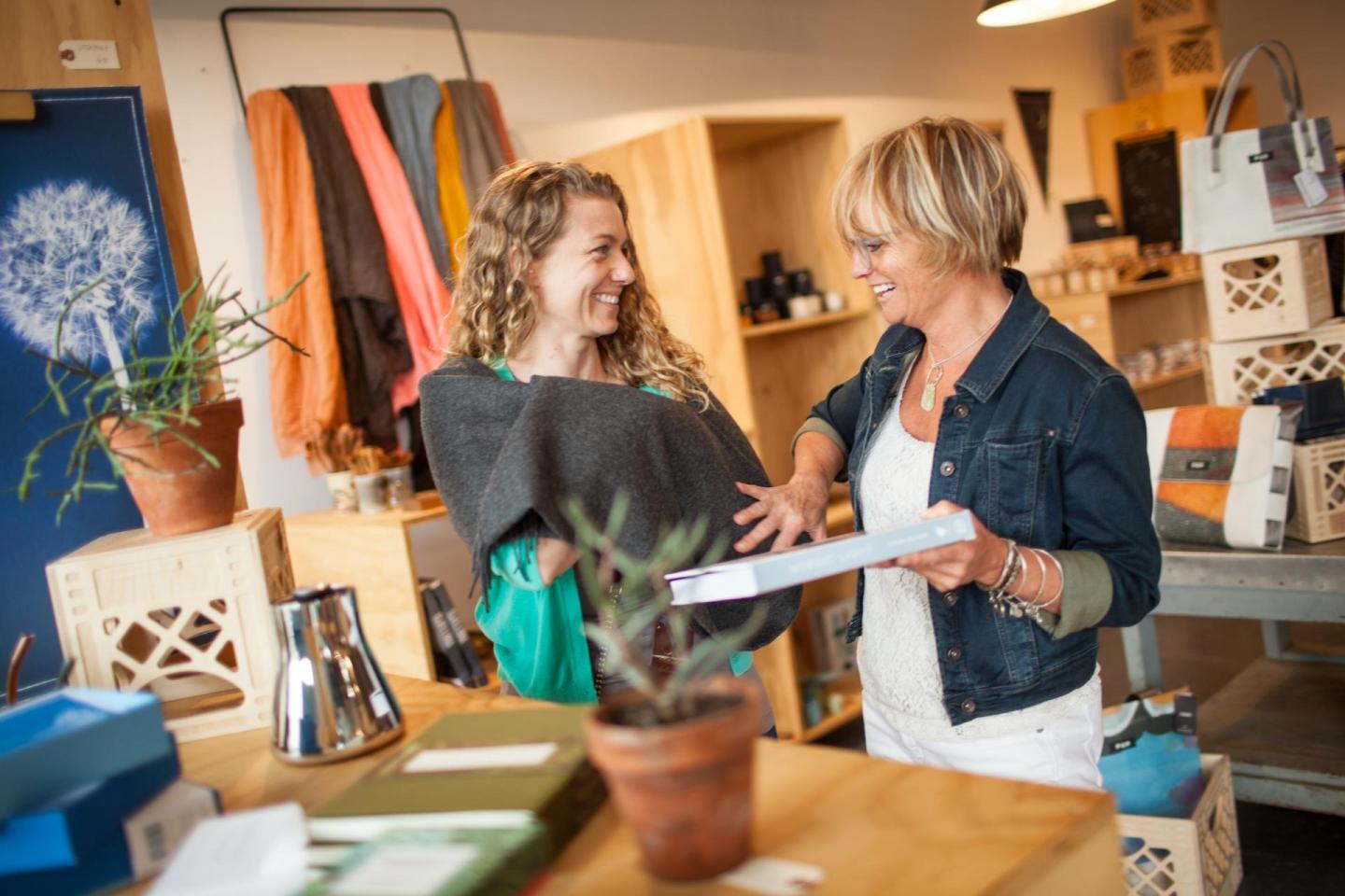 Two women smiling and discussing papers in a cozy shop with wooden shelves.
