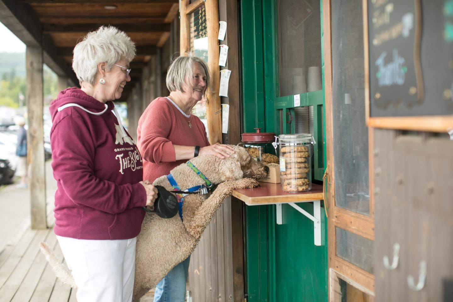Two women with a dog at an outdoor counter with a jar of treats.
