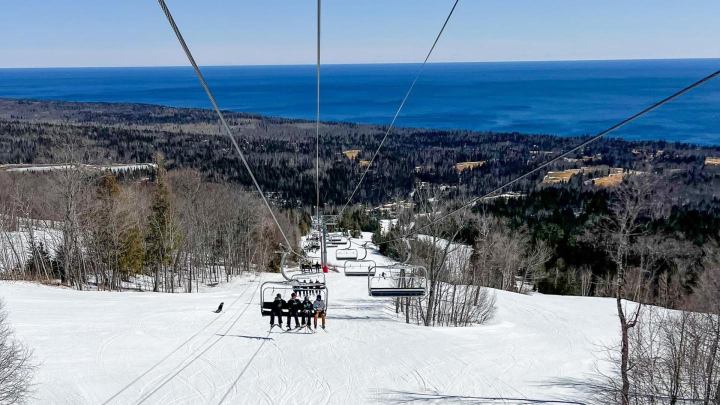 Ski lift over snowy slope with ocean view in the distance.