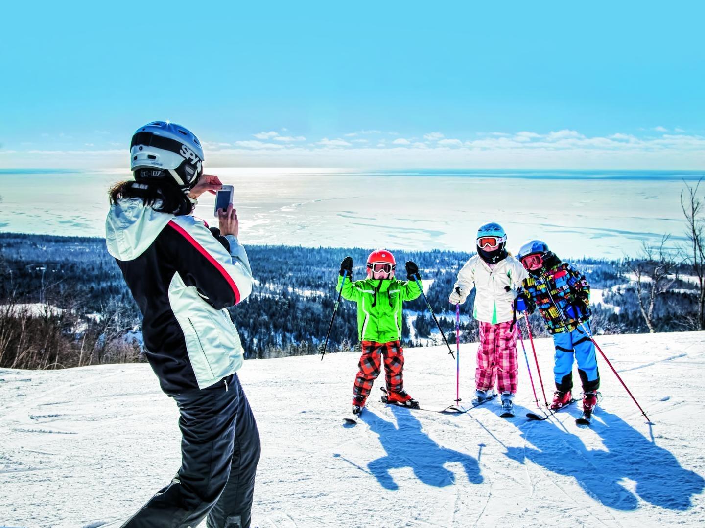 Person photographing three skiers posing on a snowy hill under a clear blue sky.