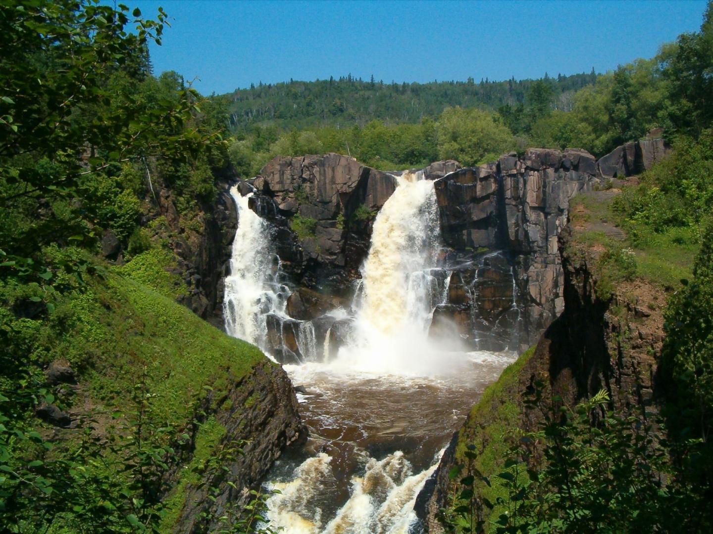 Waterfall cascading over rocky cliffs surrounded by lush green trees under a clear blue sky.