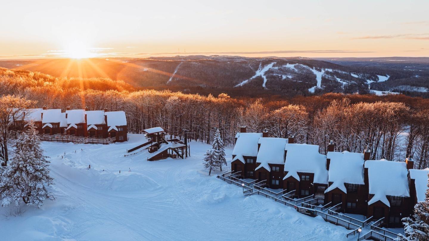 Snow-covered cabins at sunset overlooking snowy hills and trees.
