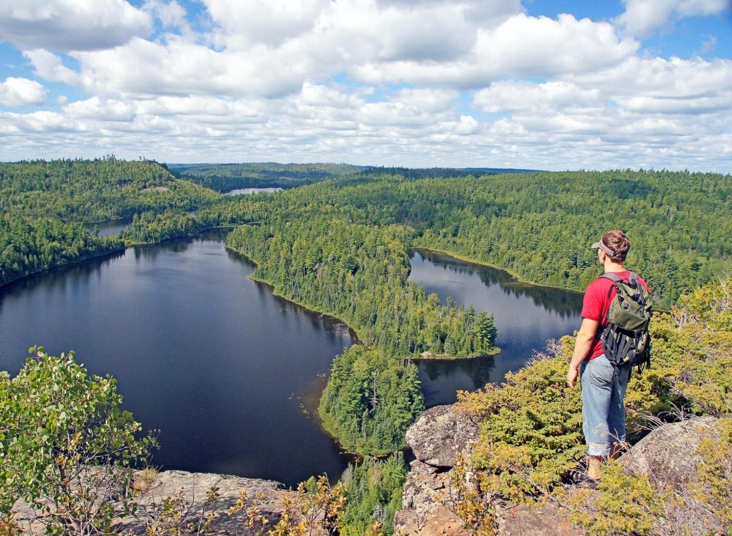 Hiker with backpack overlooks forested lakes from a rocky cliff.