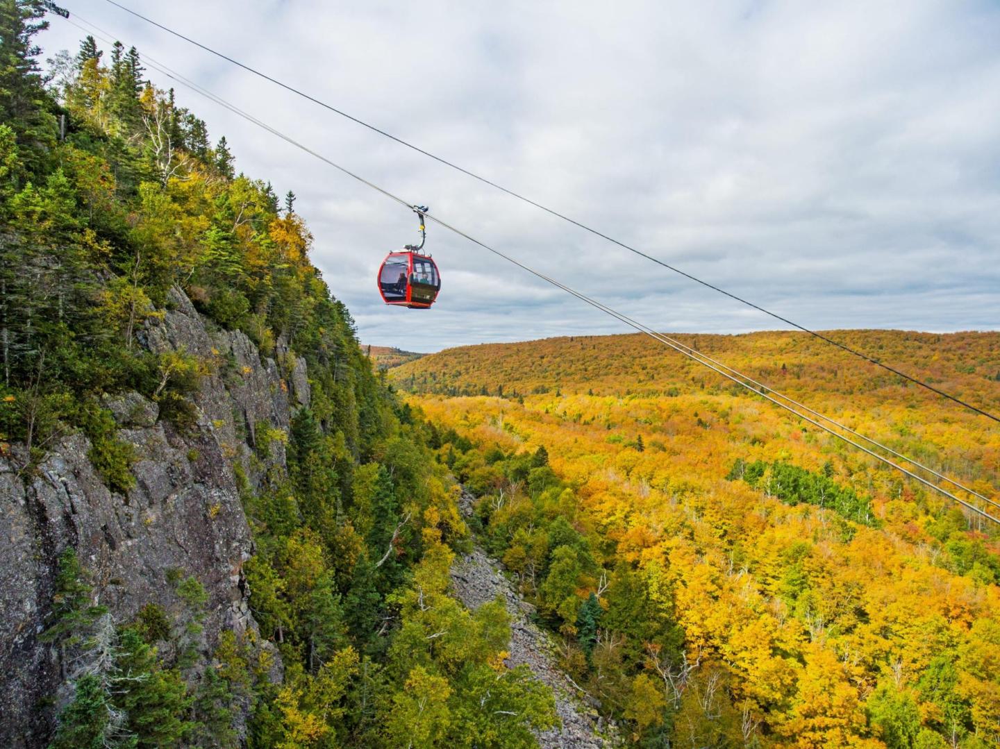 Red cable car over autumn forest and rocky cliffs under a cloudy sky.