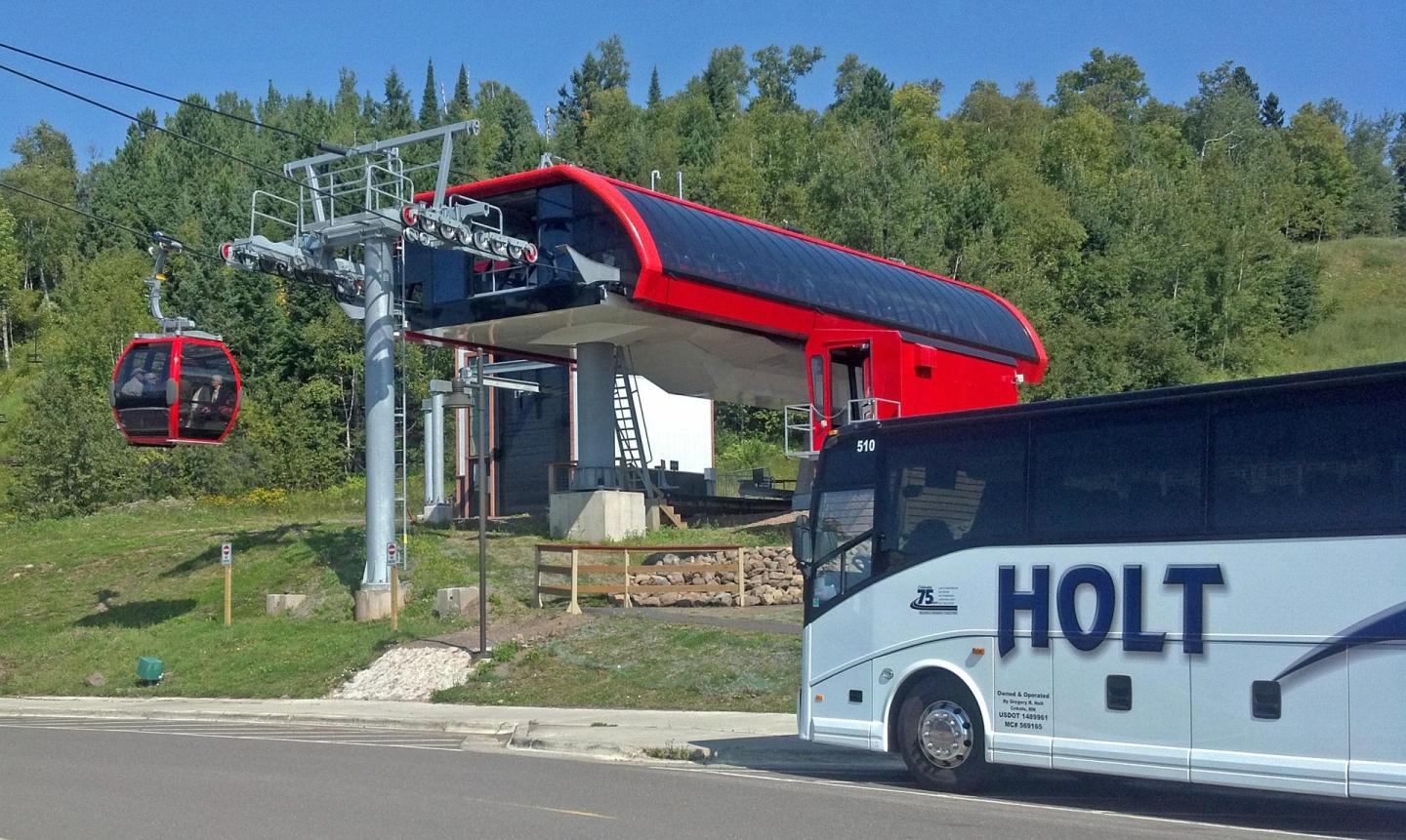 Red gondola lift and a bus in front of a lush green hillside, clear blue sky above.