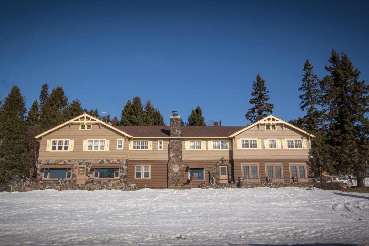 Large lodge with stone accents, snow-covered front, surrounded by tall pine trees.