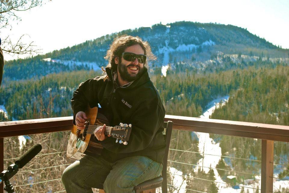 Man playing a guitar outdoors with mountains and trees in the background.