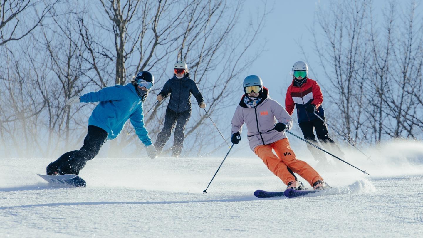 Skiers and a snowboarder on a snowy slope, trees in the background.
