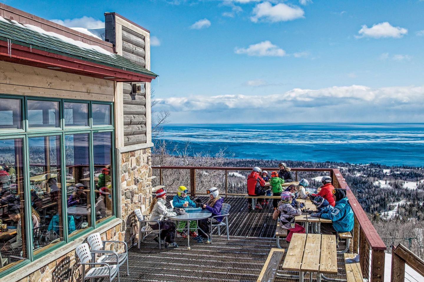 Outdoor cafe on a sunny day with people seated, overlooking a snowy landscape.
