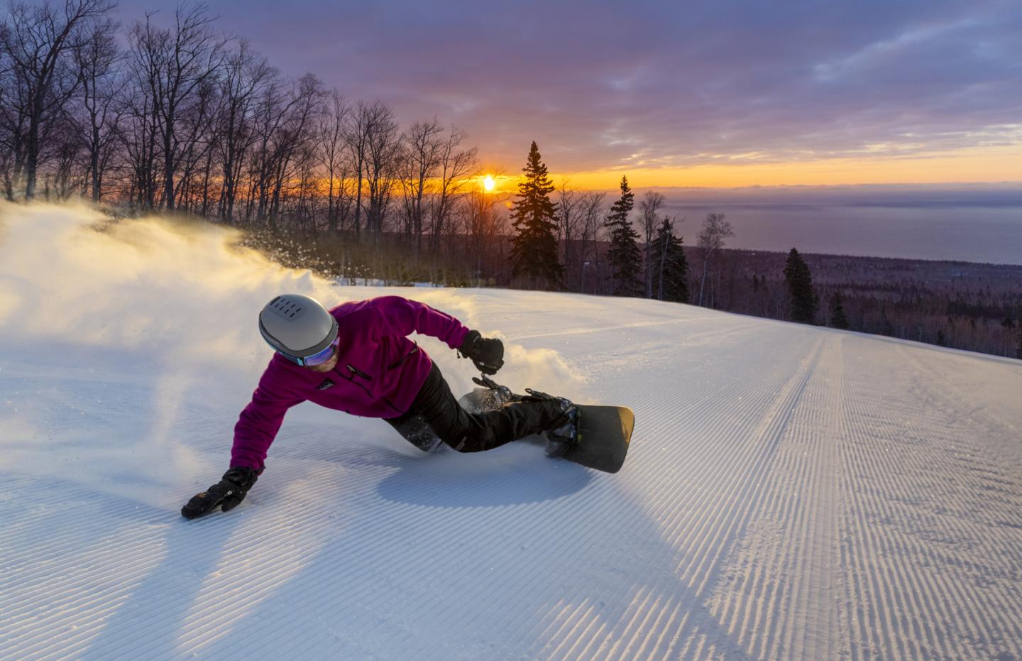 Snowboarder in a red jacket carving down a snowy slope at sunset.