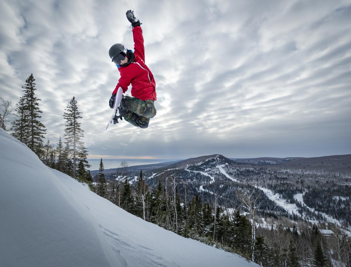 Snowboarder in red jacket jumps high on a snowy slope against a cloudy sky.