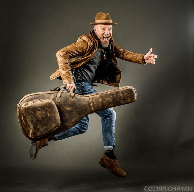 Jumping man in brown hat, leather jacket, holding guitar case, on grey background.