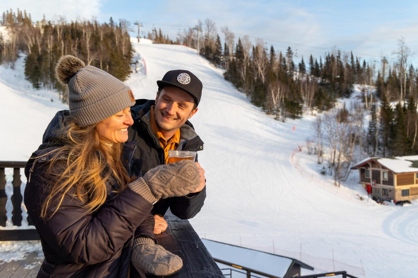 A couple enjoys drinks on a snowy ski slope, wearing winter clothes and smiling.