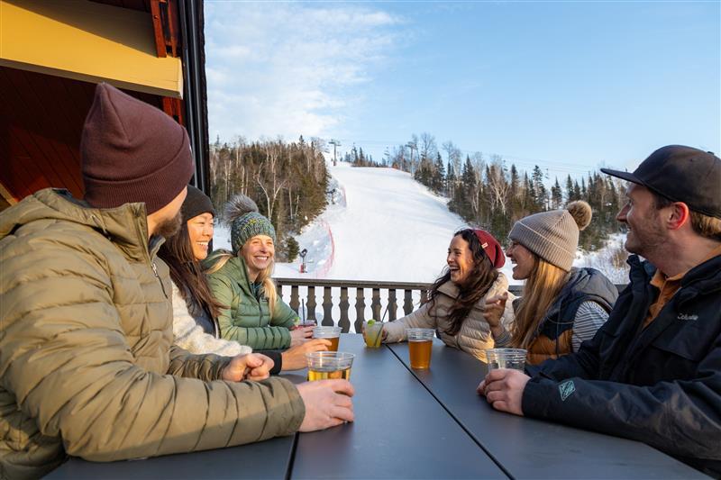 People in winter clothes enjoy drinks at a table with a snowy slope in the background.