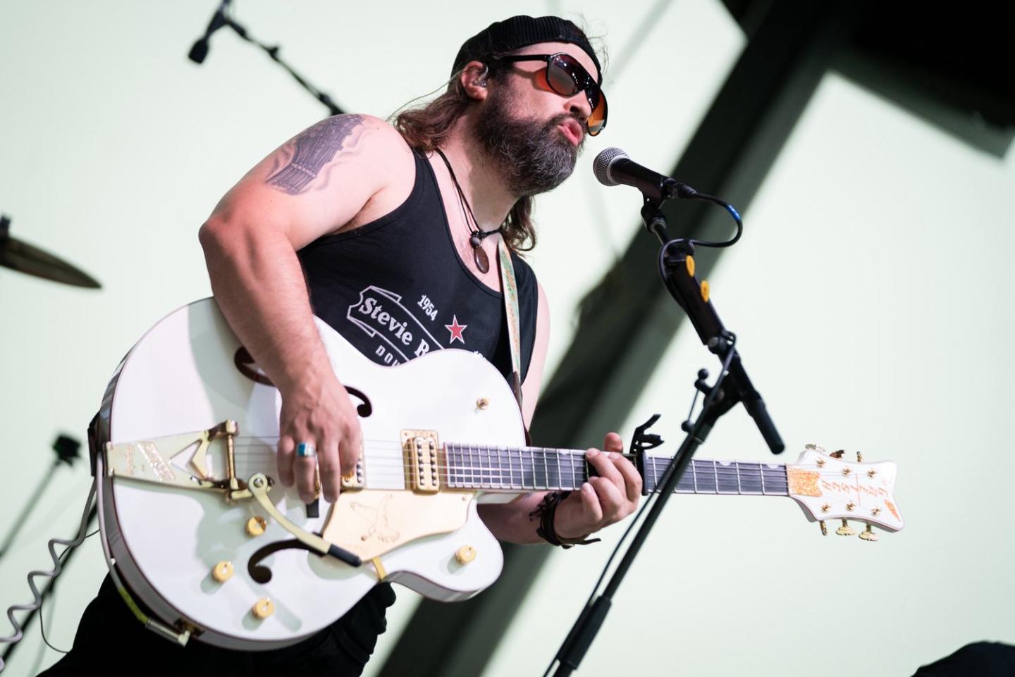 Musician plays a white electric guitar on stage, wearing sunglasses and a black tank top.